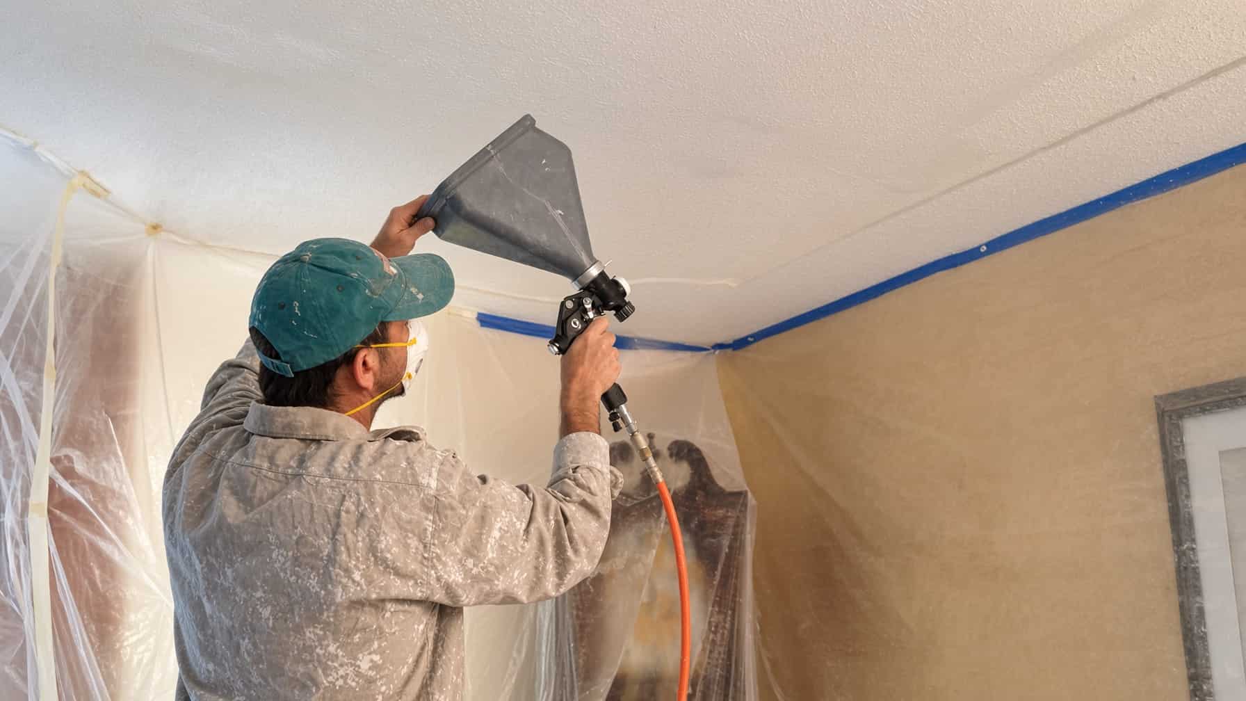 Painter spraying paint on a popcorn ceiling with the room covered in plastic in a San Diego home